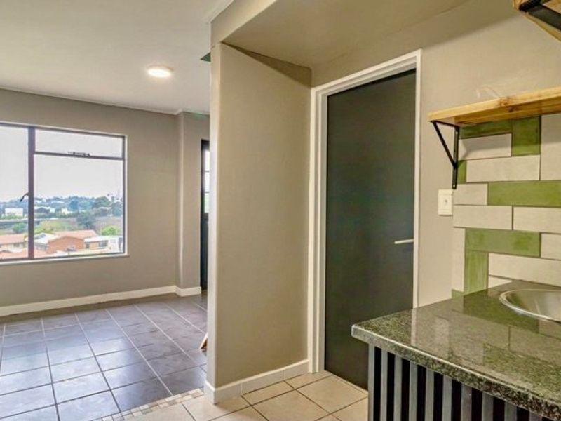 A compact kitchen area featuring granite countertops, a stainless steel sink, and decorative wall tiles, adjacent to an open-plan living space with large windows.
