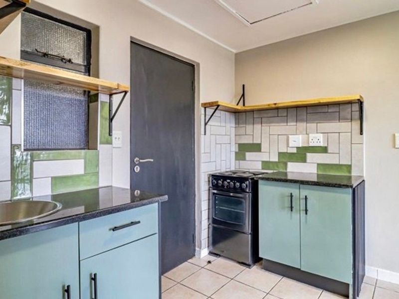 A compact kitchen featuring light green cabinetry, granite countertops, a stainless steel sink, and a tiled backsplash with floating wooden shelves.