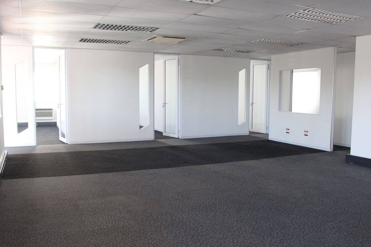 A spacious, vacant commercial office area featuring neutral partition walls, glass-paneled doors, recessed ceiling lighting, and dark grey carpeting throughout.