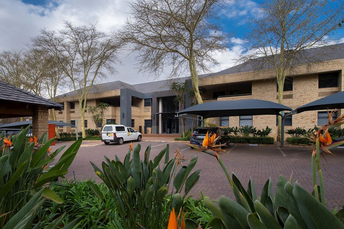 Front view of a commercial office building featuring a brick facade, paved parking area with shade cloth carports, and established garden landscaping.