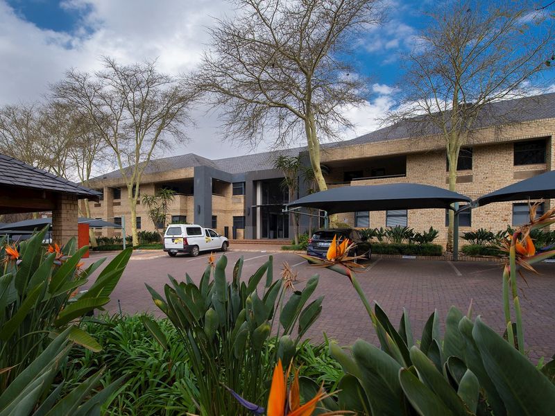 Front view of a commercial office building featuring a brick facade, paved parking area with shade cloth carports, and established garden landscaping.