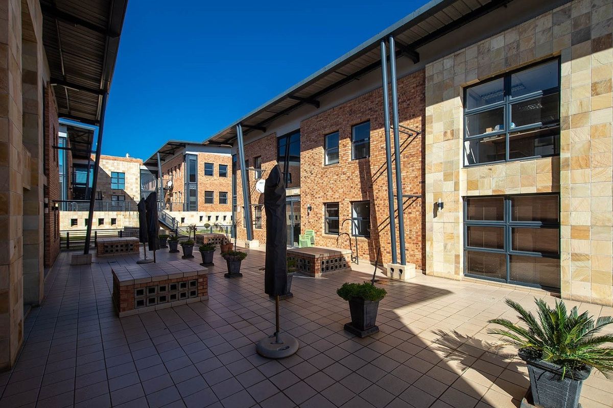 A spacious outdoor patio area featuring tiled flooring, brick and stone facade finishes, and modern metal structural supports within a commercial office complex.