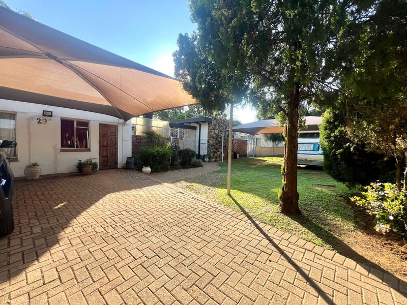 Front exterior view featuring a paved driveway, shade sail covering, and established garden area with mature trees.