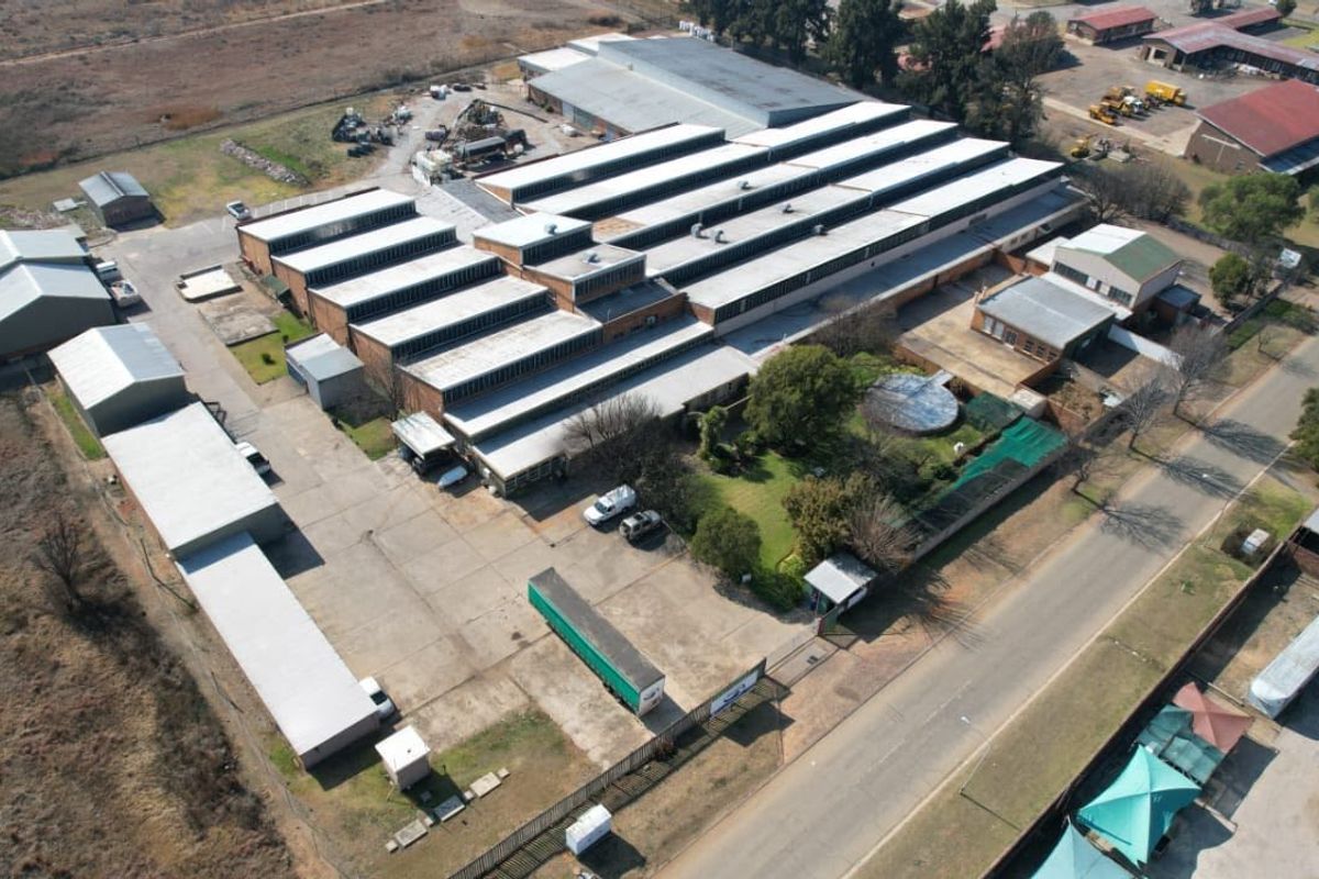 Aerial view of a large industrial warehouse facility featuring multiple sawtooth roof sections, extensive concrete yard space, and perimeter security fencing.