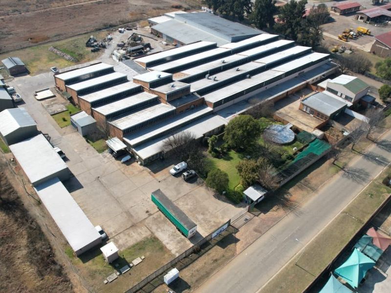 Aerial view of a large industrial warehouse facility featuring multiple sawtooth roof sections, extensive concrete yard space, and perimeter security fencing.