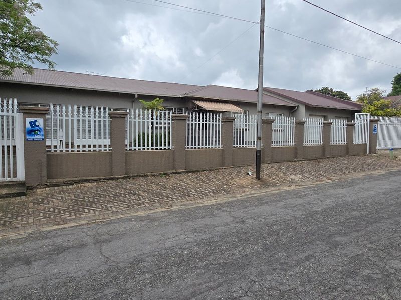 Front exterior view of a single-story house featuring a perimeter wall with security fencing, paved sidewalk, and gated access.