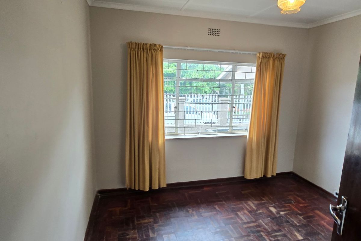 A bright, empty bedroom featuring classic parquet flooring, a window with security bars, and neutral wall finishes.