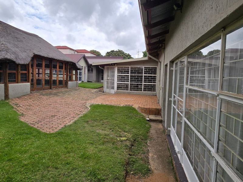 Rear exterior view featuring a thatched roof structure, paved patio area, and established lawn with metal-framed windows and security bars.