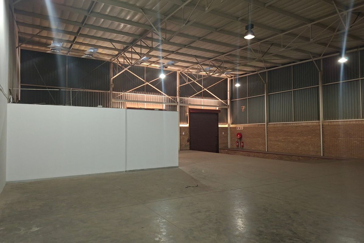 Spacious industrial warehouse unit featuring high ceilings with exposed steel trusses, concrete flooring, and a large roller shutter door for easy access.