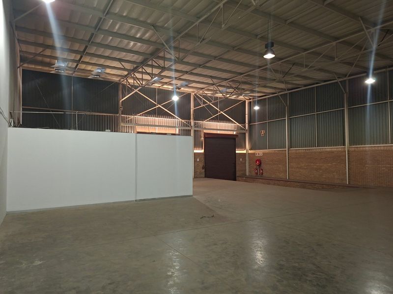 Spacious industrial warehouse unit featuring high ceilings with exposed steel trusses, concrete flooring, and a large roller shutter door for easy access.