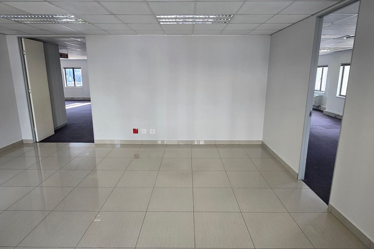 An empty, well-lit commercial office space featuring neutral tiled flooring, a suspended ceiling with recessed lighting, and multiple access points to adjacent areas.