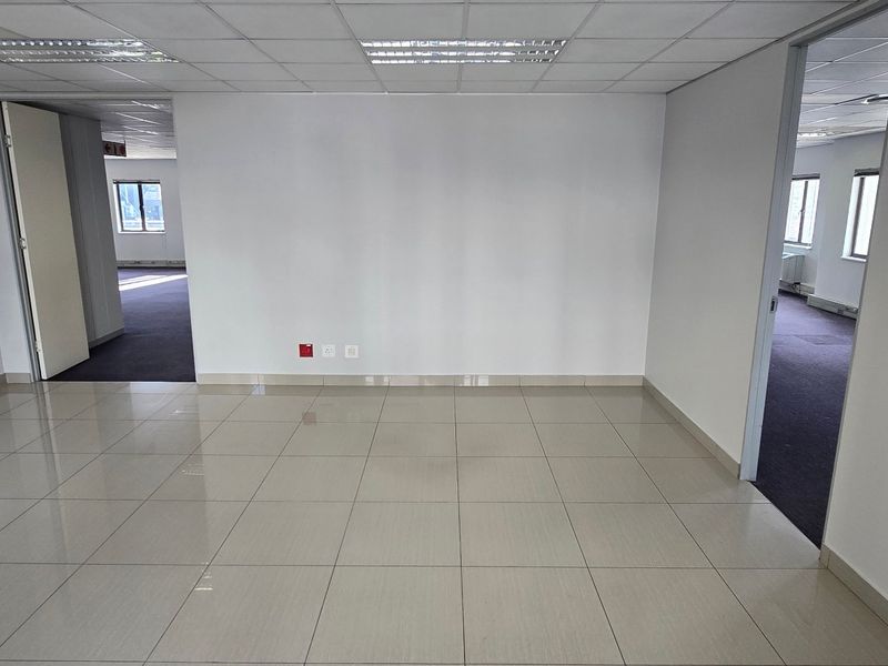 An empty, well-lit commercial office space featuring neutral tiled flooring, a suspended ceiling with recessed lighting, and multiple access points to adjacent areas.