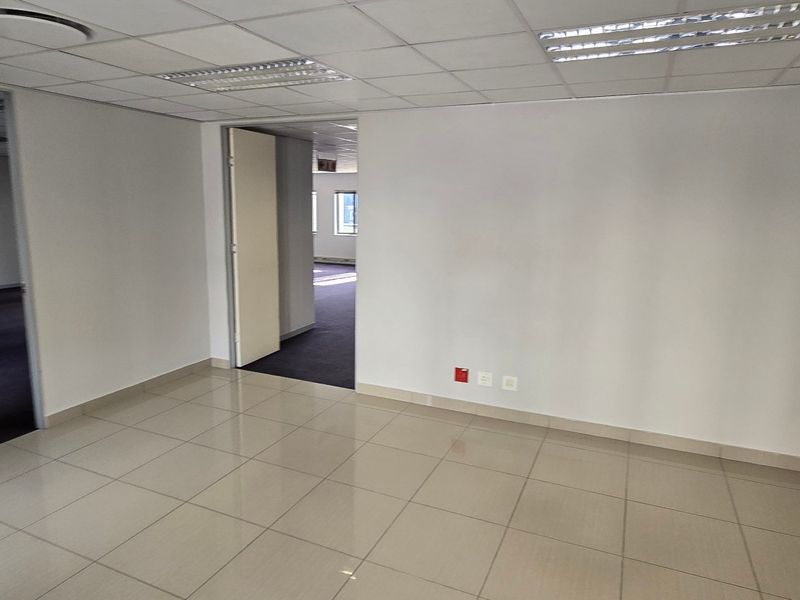 An empty, well-lit commercial office space featuring tiled flooring, a suspended ceiling with recessed lighting, and multiple access points to adjacent office areas.