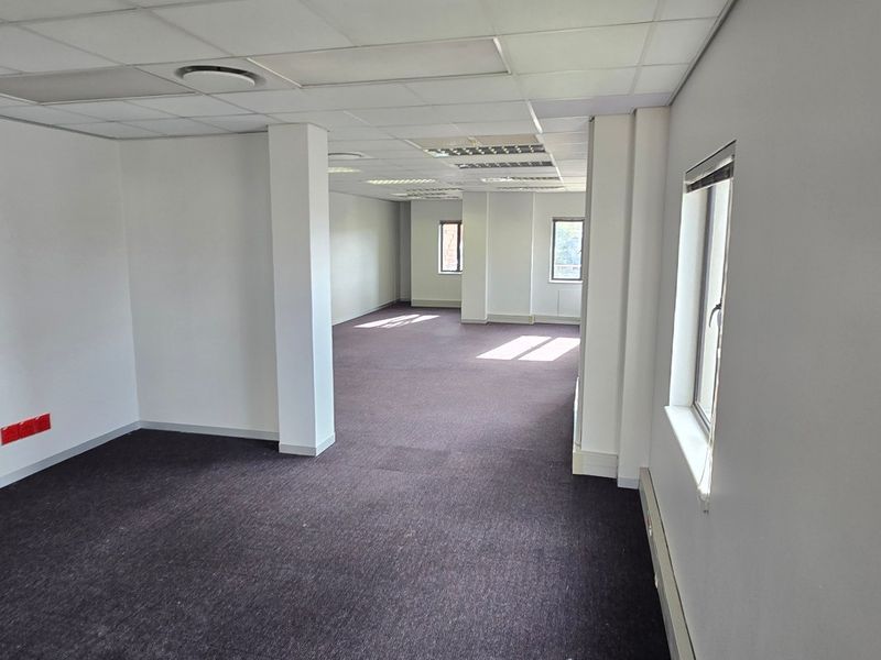 A spacious, vacant commercial office area featuring neutral walls, carpeted flooring, and a suspended ceiling with integrated lighting.