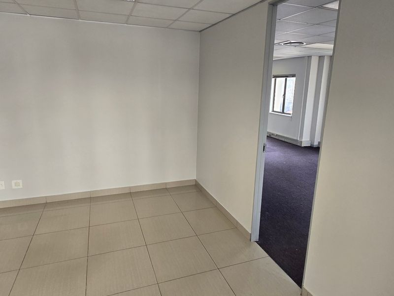 An empty commercial office space featuring tiled flooring, a suspended ceiling, and integrated electrical and data connectivity, leading into an adjacent carpeted office area.