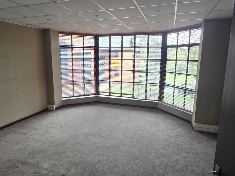 An empty office space featuring a large bay window, suspended ceiling with recessed lighting, and fitted carpeting.