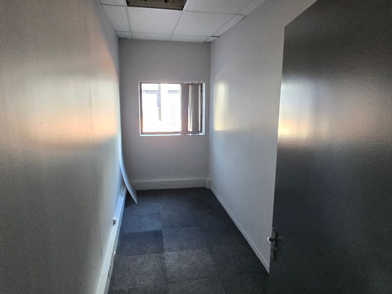 A small, empty office space featuring carpet tiles, a suspended ceiling, and a window with vertical blinds.