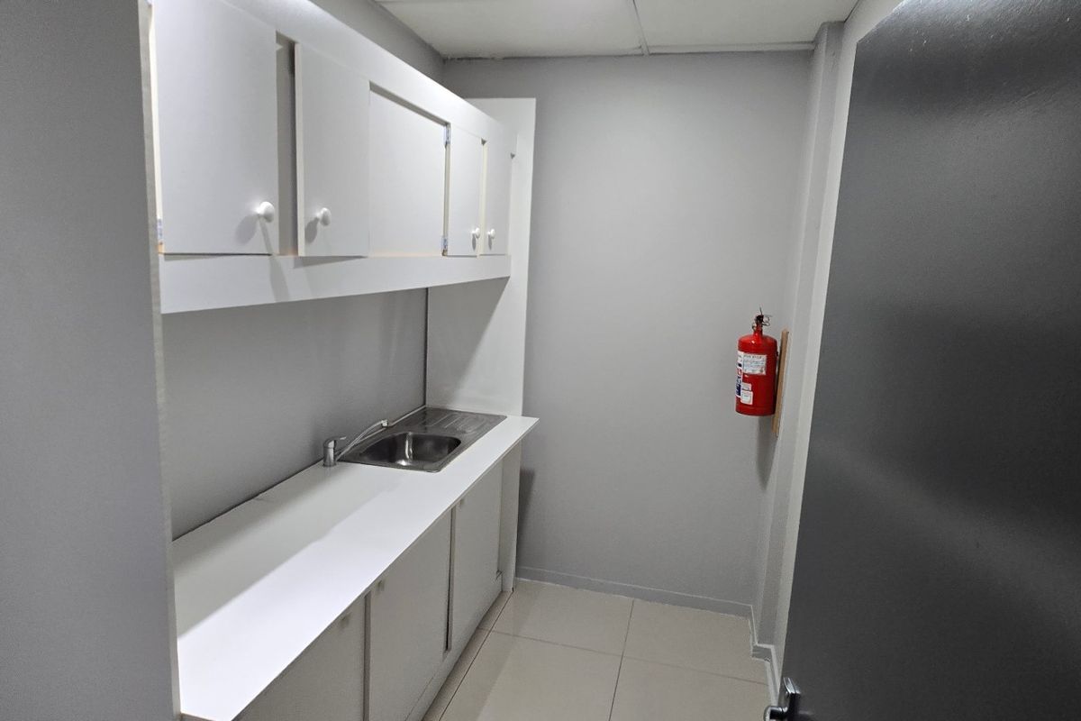 A compact commercial kitchenette featuring built-in cabinetry, a stainless steel sink, and tiled flooring, suitable for office or utility use.