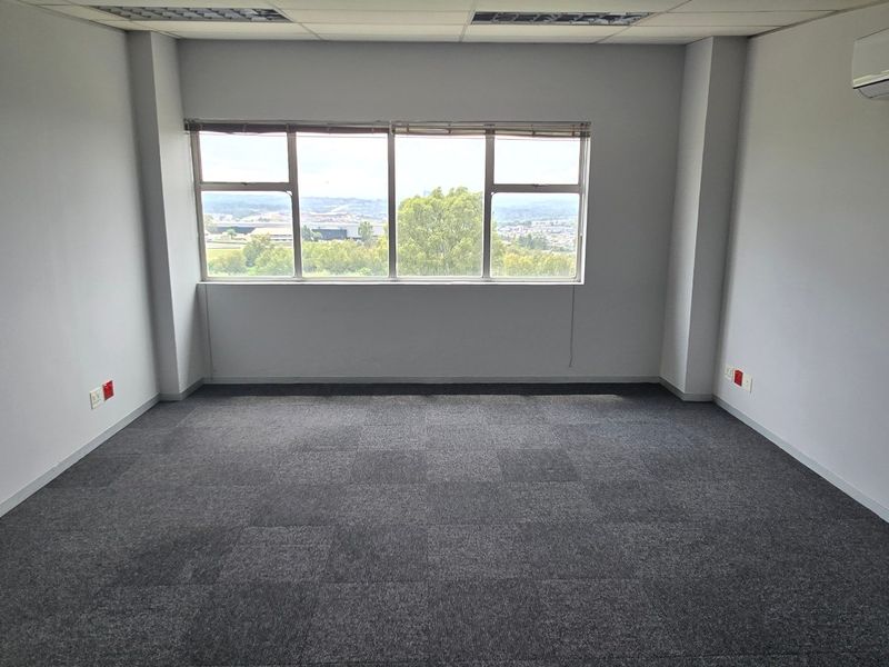 A bright, empty office space featuring carpeted flooring, a suspended ceiling with recessed lighting, and a wall-mounted air conditioning unit.