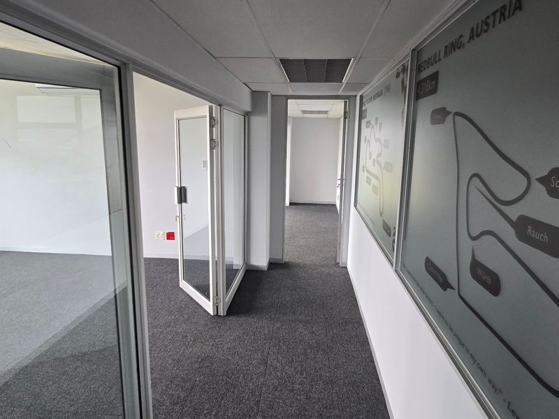 A modern commercial hallway featuring glass partition walls, carpeted flooring, and a suspended ceiling, providing a clean and professional office layout.