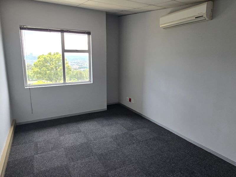 A clean, empty office space featuring carpet tiles, a suspended ceiling, and a wall-mounted air conditioning unit with a view through the window.