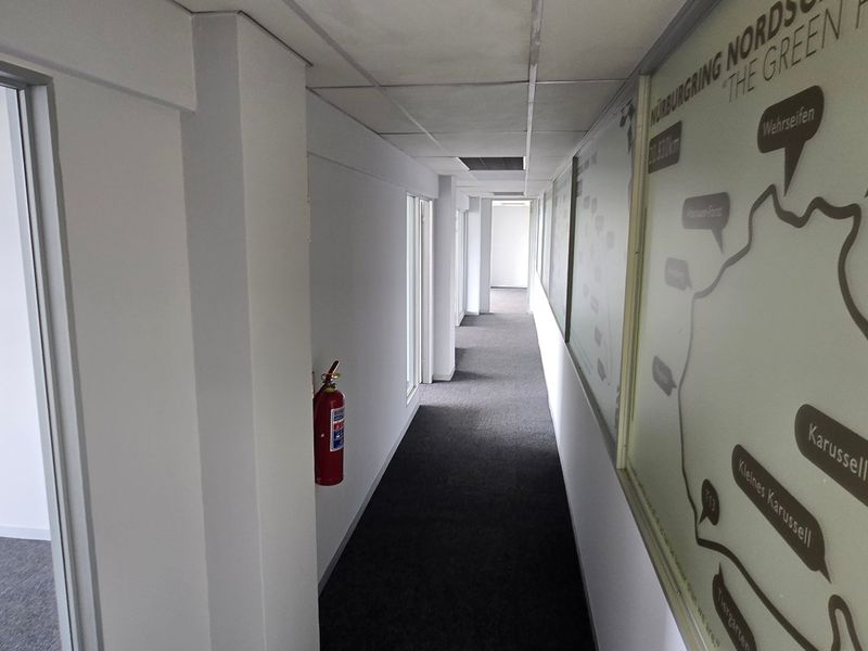 A clean, well-lit commercial hallway featuring carpeted flooring, a suspended ceiling, and glass partition walls leading to adjacent office spaces.