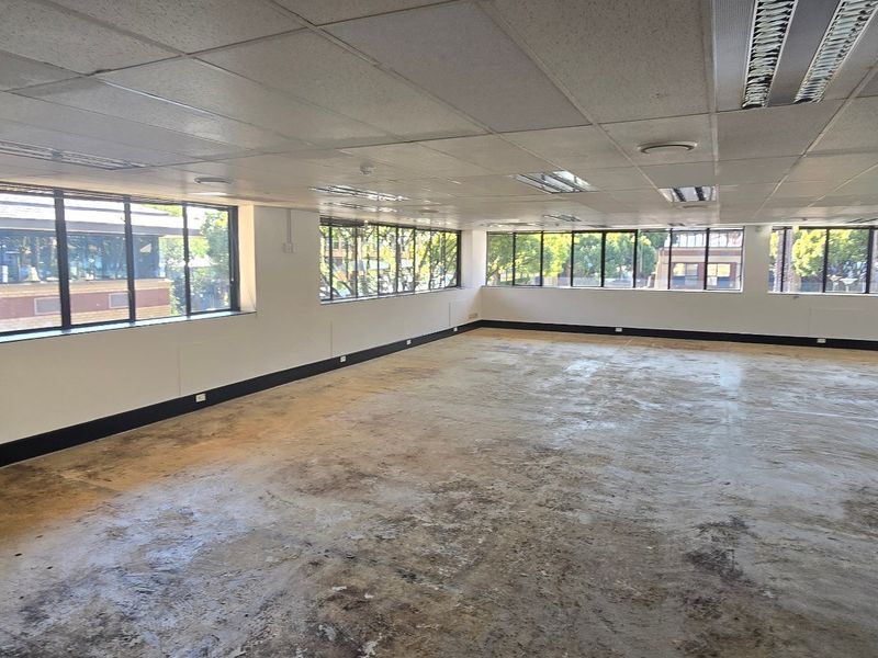 A spacious, open-plan commercial office area featuring large windows, suspended ceilings with recessed lighting, and exposed concrete flooring ready for refurbishment.