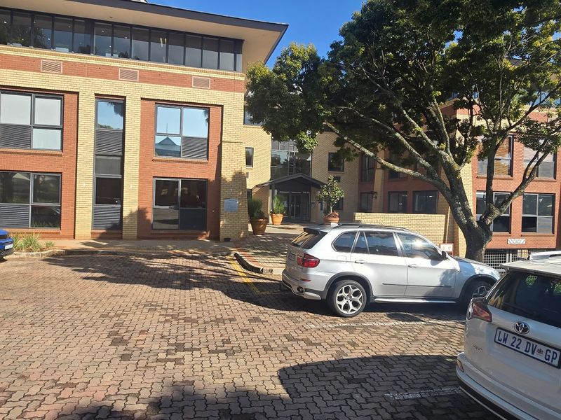 A multi-story commercial office building featuring a brick facade, large glass windows, and a paved parking area at the front entrance.