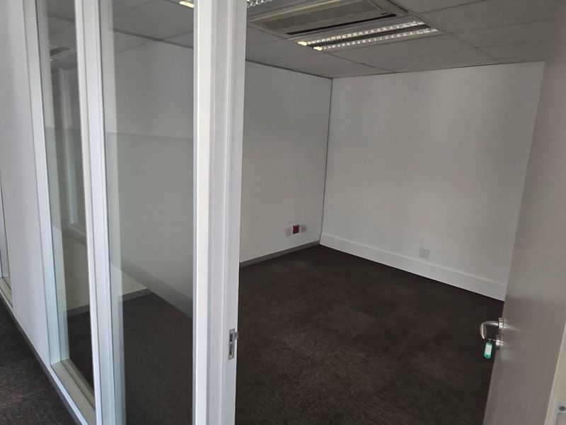 A vacant, modern office space featuring glass partition walls, carpeted flooring, and integrated ceiling lighting, suitable for a professional workspace.
