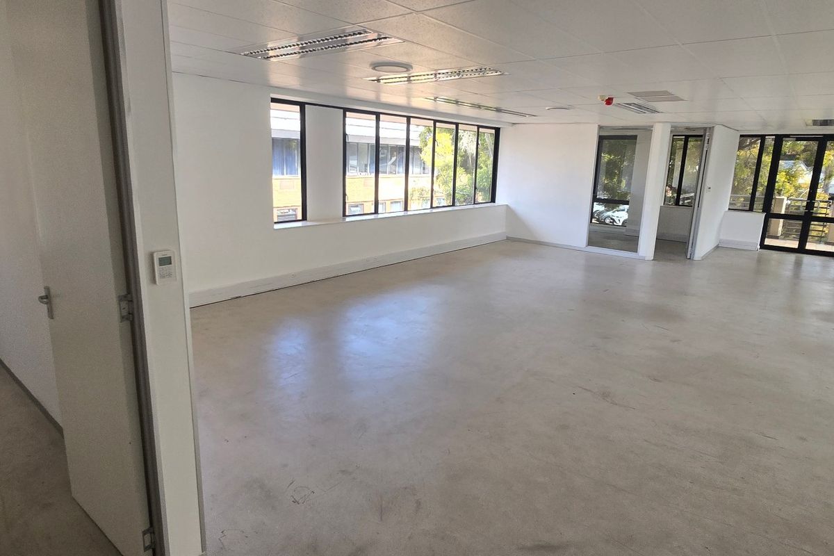 A spacious, vacant commercial office area featuring large windows, concrete flooring, and a suspended ceiling with integrated lighting, offering a versatile open-plan workspace.