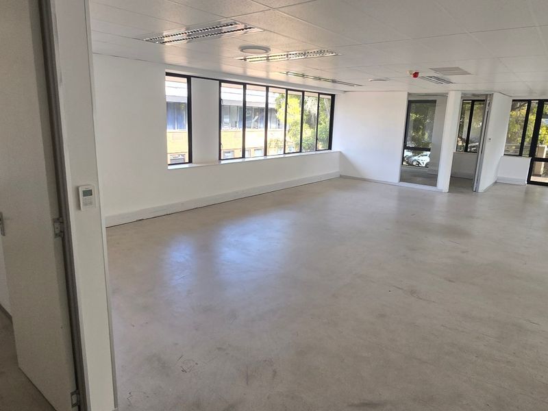A spacious, vacant commercial office area featuring large windows, concrete flooring, and a suspended ceiling with integrated lighting, offering a versatile open-plan workspace.