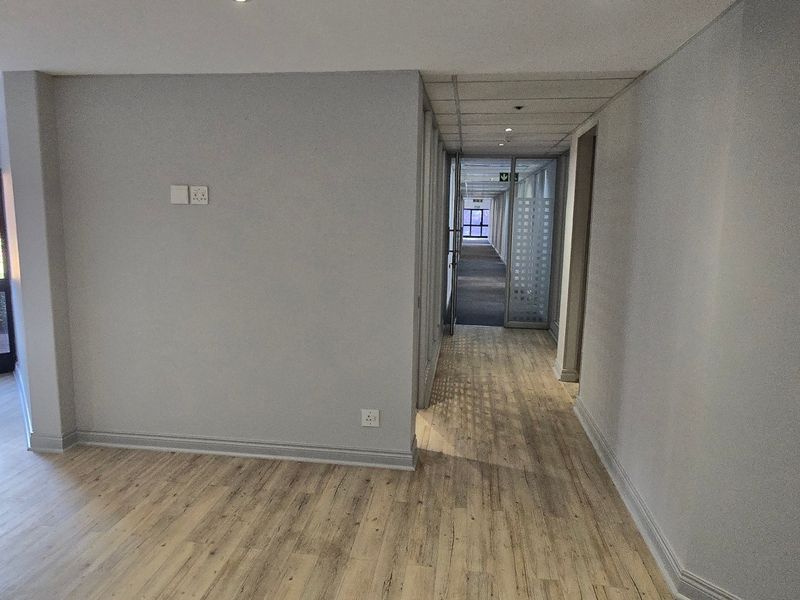 A bright, empty commercial hallway featuring modern laminate flooring, recessed lighting, and glass partition doors leading to an adjacent office corridor.
