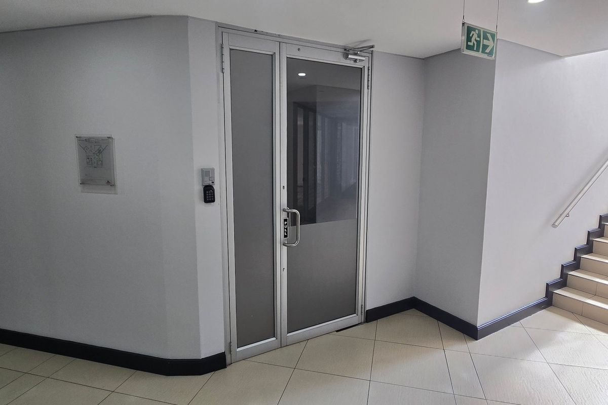 A clean commercial hallway featuring tiled flooring, a secure aluminium glass door with keypad access, and a staircase with a handrail.