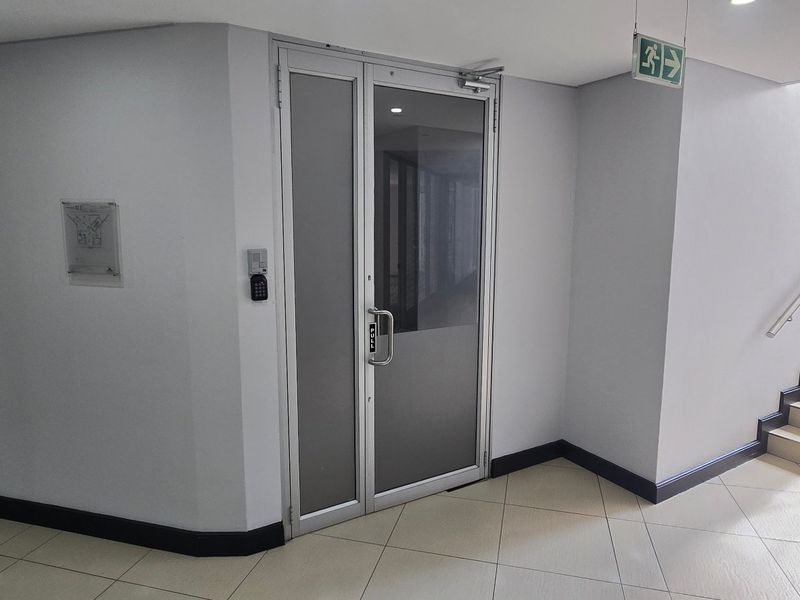 A clean commercial hallway featuring tiled flooring, a secure aluminium glass door with keypad access, and a staircase with a handrail.
