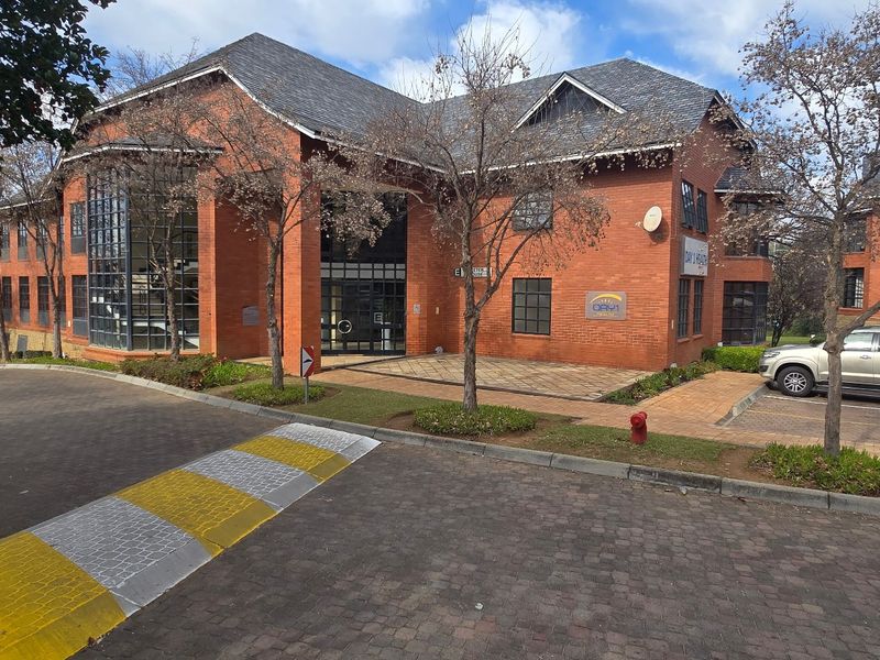 Front exterior view of a multi-story commercial office building featuring a red brick facade, pitched roof, and paved parking area.