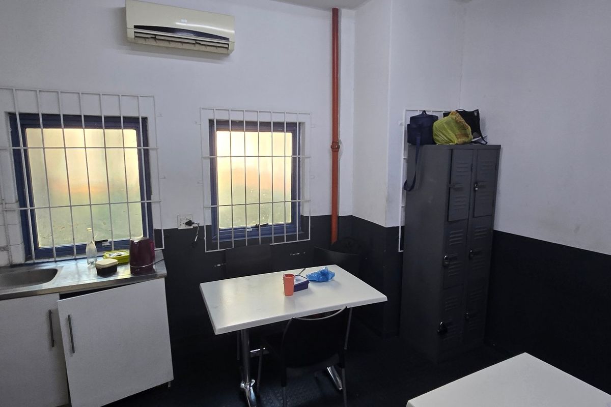 A functional industrial kitchen area featuring a stainless steel sink, built-in cabinetry, security window bars, and a wall-mounted air conditioning unit.