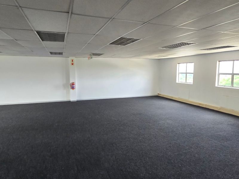 A spacious, open-plan commercial office area featuring neutral walls, dark carpet tiles, and a suspended ceiling with integrated lighting.