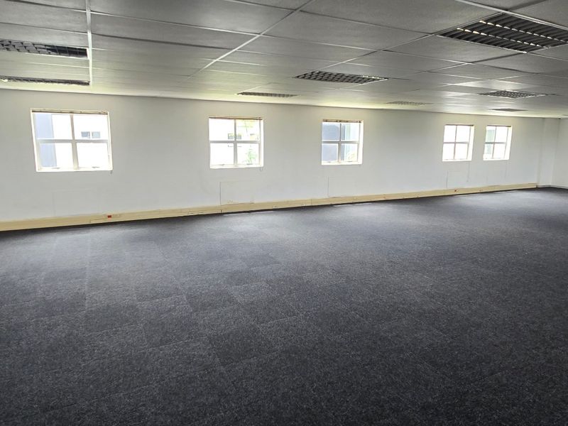 A spacious, open-plan commercial office area featuring dark carpet tiles, a suspended ceiling with recessed lighting, and multiple windows providing natural light.
