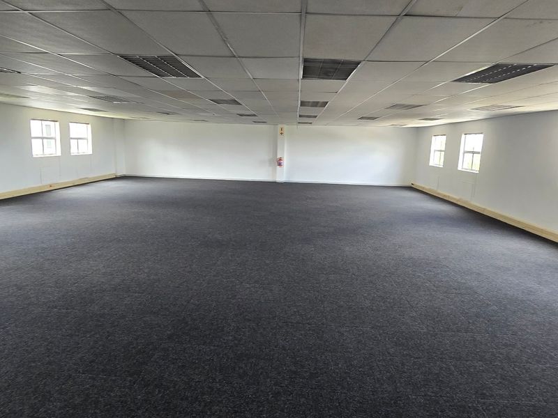 A spacious, empty open-plan commercial office area featuring a suspended ceiling, recessed lighting, and dark carpeting throughout.