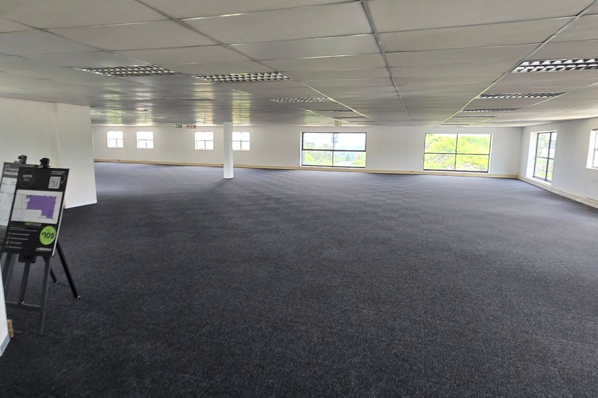 A spacious, open-plan commercial office area featuring a suspended ceiling, recessed lighting, and dark carpet tiles, offering a versatile workspace with ample natural light.