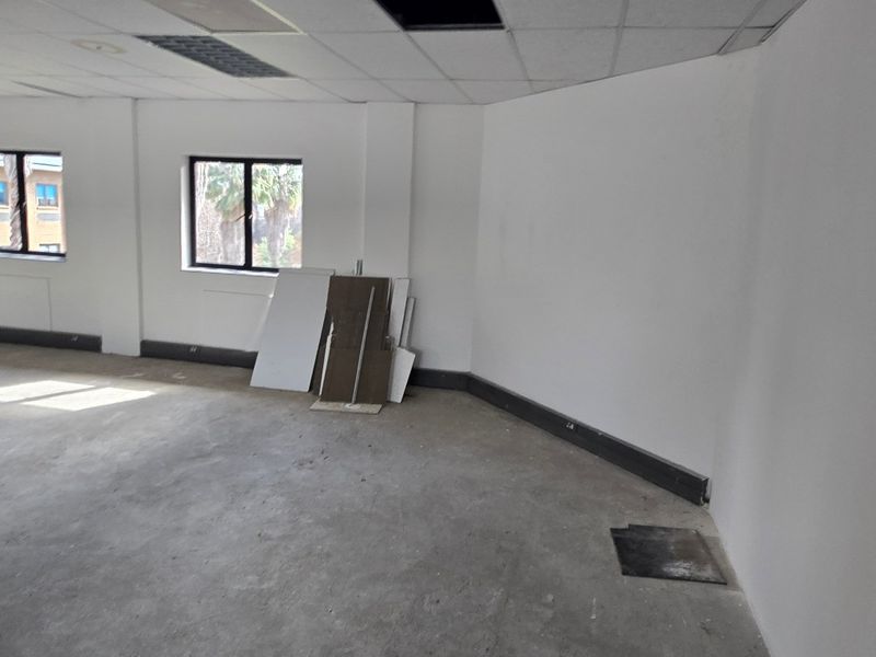 An empty commercial office space featuring a suspended ceiling, recessed lighting, and large windows, currently requiring flooring and ceiling tile completion.