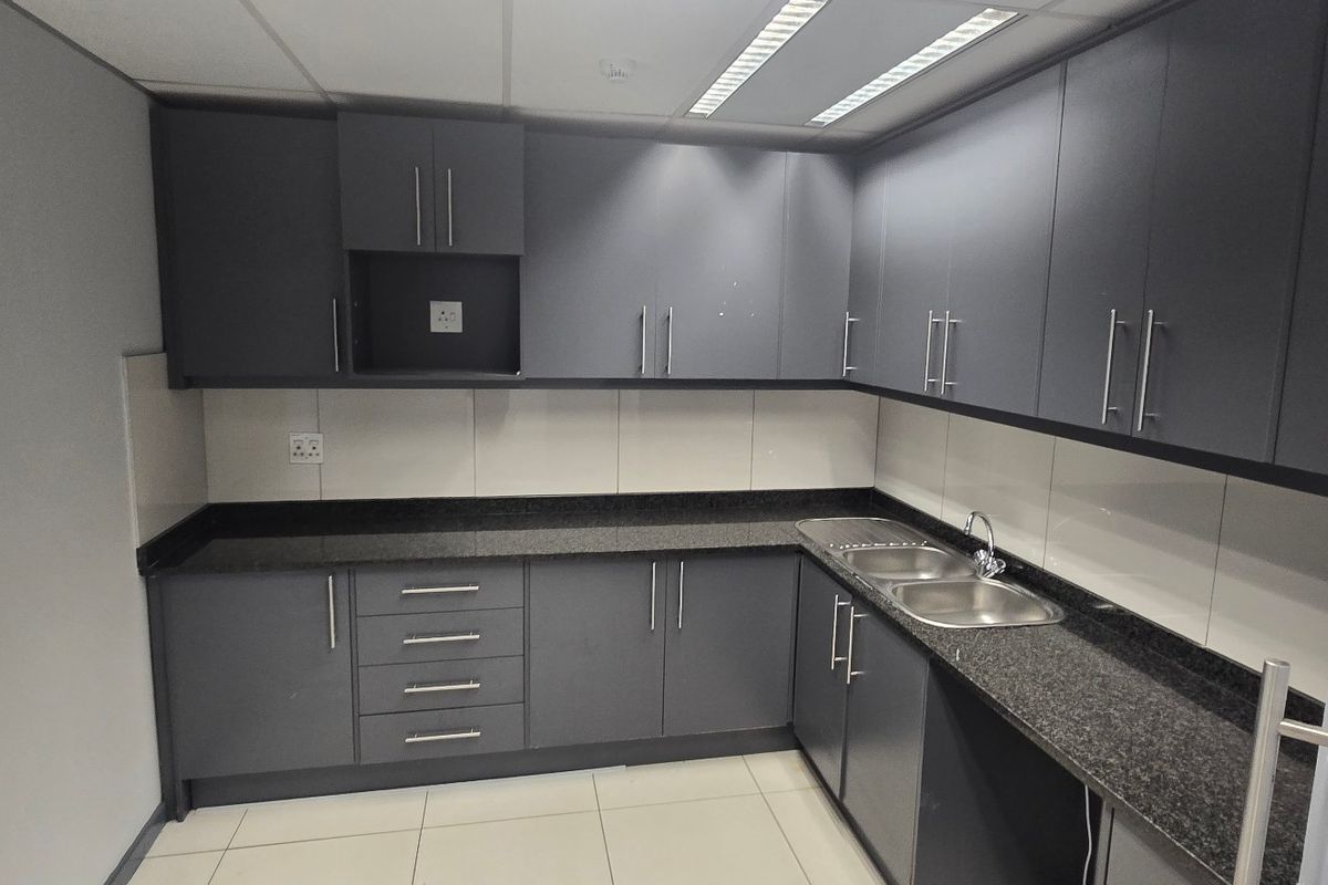 A clean and functional commercial kitchenette featuring modern dark grey cabinetry, granite countertops, a double stainless steel sink, and neutral tiled finishes.
