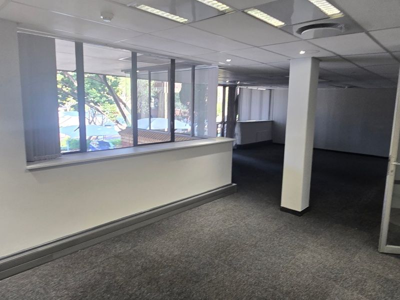 An open-plan commercial office space featuring carpeted flooring, a suspended ceiling with recessed lighting, and large windows providing natural light.