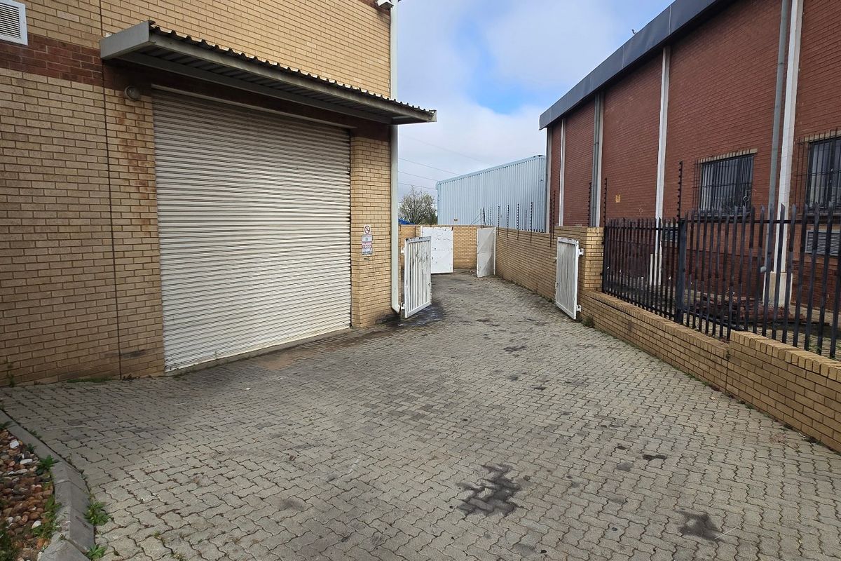 Rear exterior area of a commercial property featuring a roller shutter door, paved access path, and secure brick boundary wall with electric fencing.