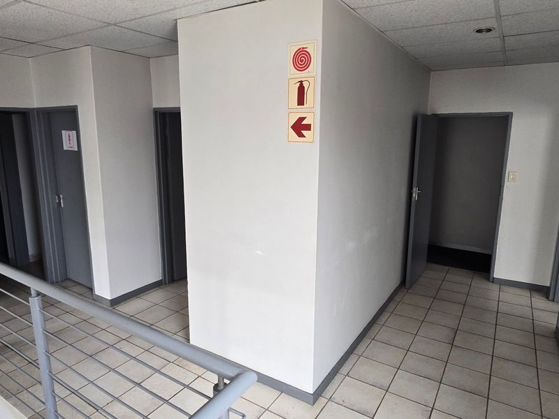 A functional commercial hallway featuring ceramic tiled flooring, a suspended ceiling, and a metal safety balustrade leading to multiple office or utility doorways.