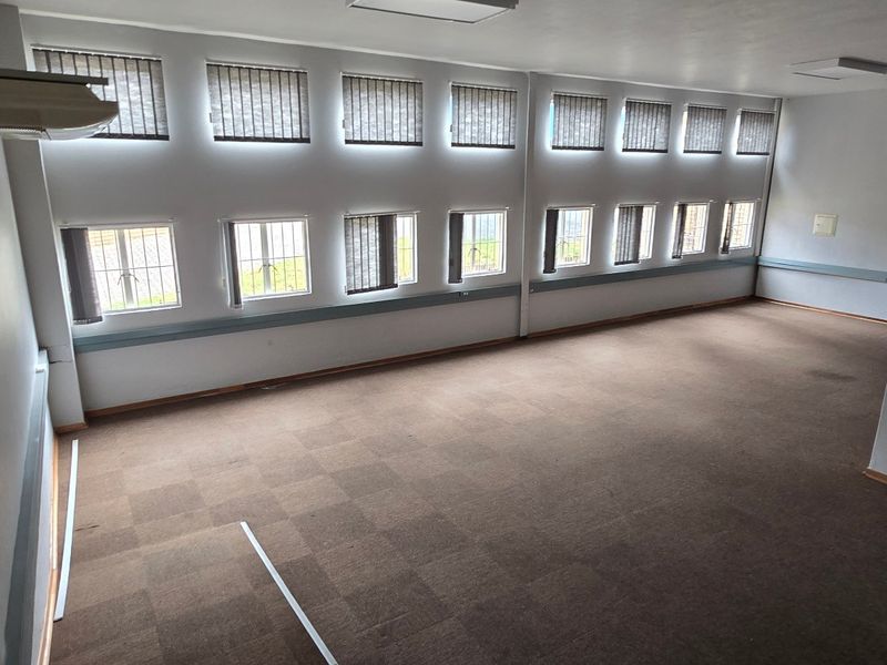 Spacious, empty commercial office area featuring carpeted flooring, multiple windows with security bars, vertical blinds, and an integrated air conditioning unit.
