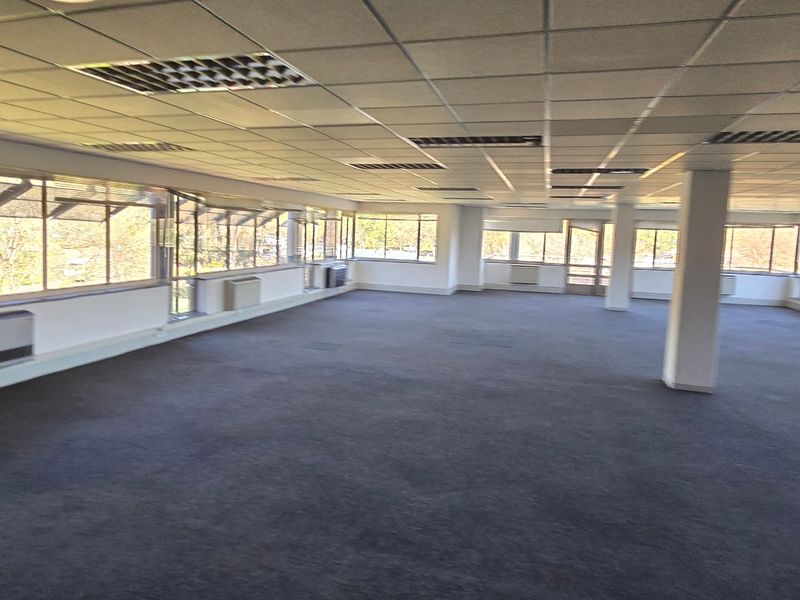 Spacious open-plan commercial office area featuring a suspended ceiling, large windows providing natural light, and perimeter air conditioning units.