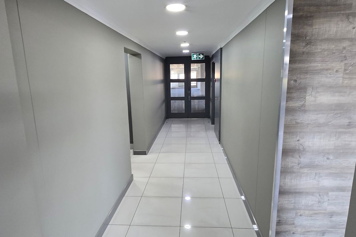 A clean, modern commercial hallway featuring polished ceramic floor tiles, recessed downlighting, and a wood-accented wall leading to a glass-panelled exit door.