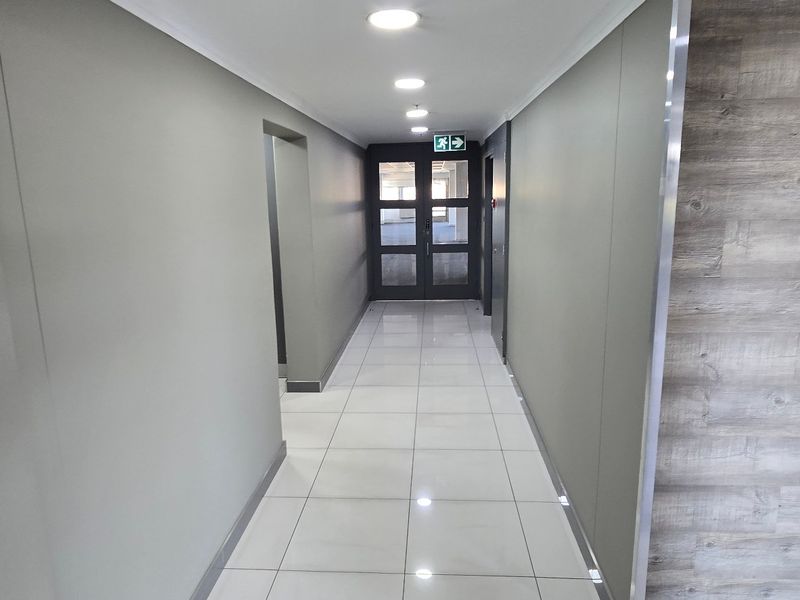 A clean, modern commercial hallway featuring polished ceramic floor tiles, recessed downlighting, and a wood-accented wall leading to a glass-panelled exit door.