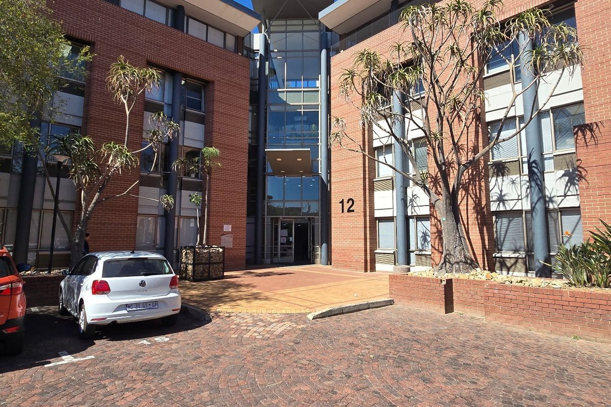 Front elevation of a multi-story commercial office building featuring a brick facade, glass entrance atrium, and paved parking area.